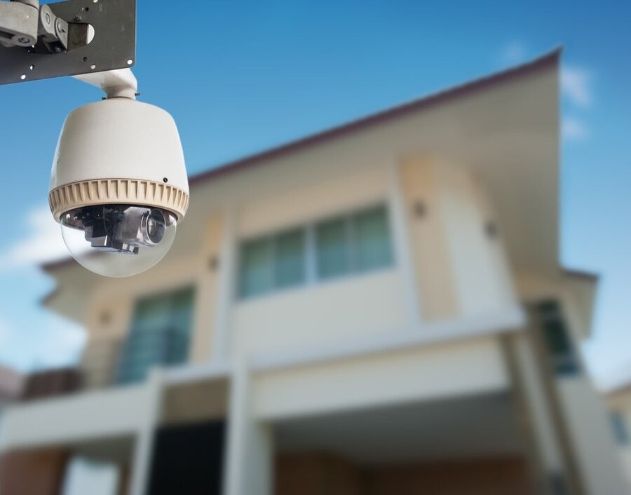 A smart surveillance camera in focus in the foreground with a home’s exterior in the background.