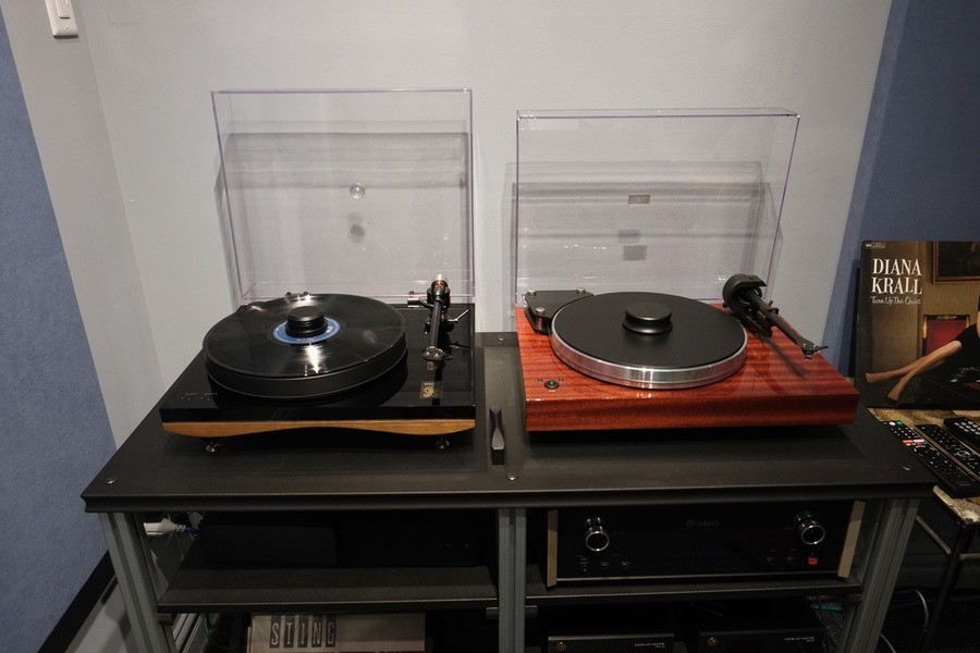 Two premium turntables sit side by side on a sleek audio console, one in a modern black design with a curved wooden base and the other in a rich wooden finish, both with clear dust covers lifted to display their precision tonearms.