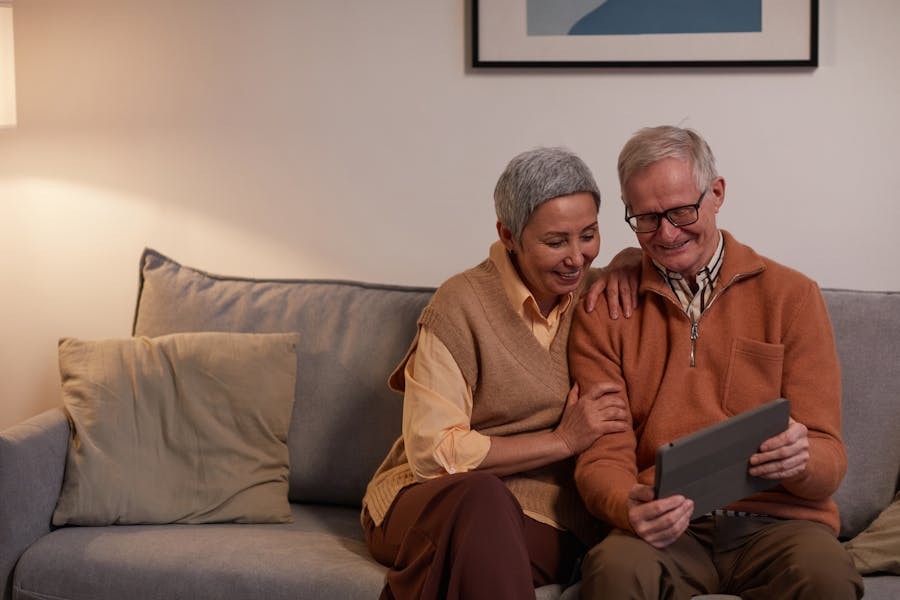 Smiling senior couple sitting on a sofa using a tablet together, enjoying the benefits of smart home technology.